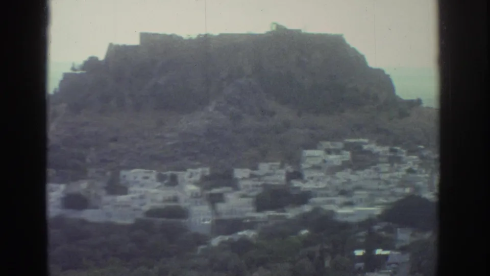 1976 Greece 8mm frame showing a whitewashed hillside village beneath a rocky ridge with the sea beyond.