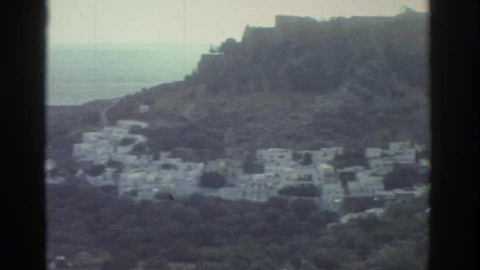 1976 Greece 8mm frame of white buildings on a rocky slope, with a narrow view toward the sea between hills.