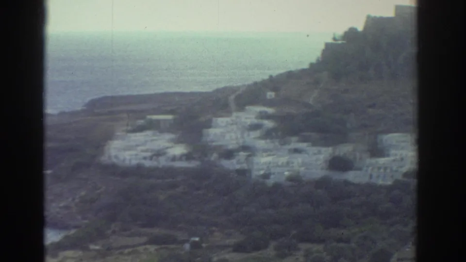 1976 Greece 8mm frame during a pan, showing sea, rocky ground, sparse greenery, and white village structures.
