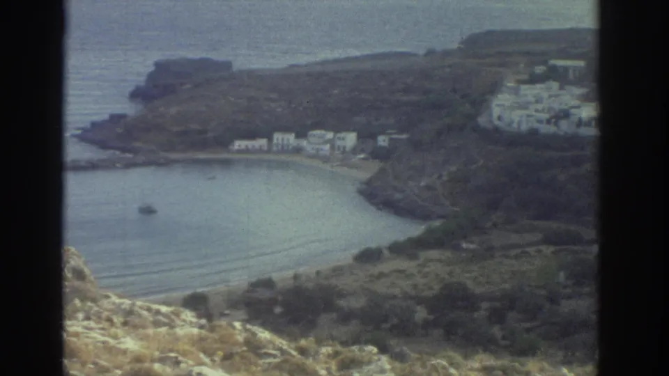 1976 Greece 8mm frame showing a quiet cove, rocky headlands, white coastal buildings, and a small boat on the water.