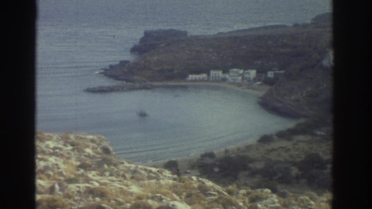 1976 Greece 8mm frame looking down from a rocky cliff toward a sheltered bay, shoreline buildings, and a small boat on calm water.