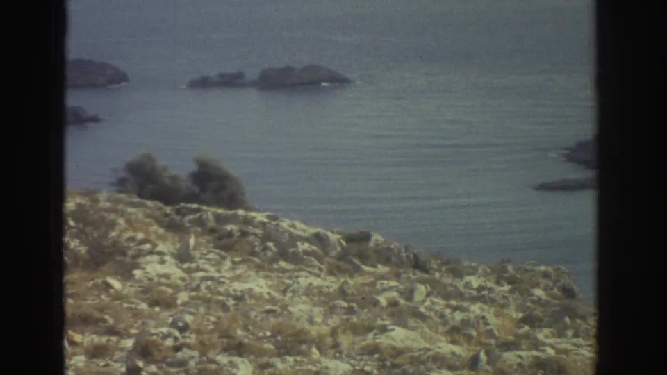 1976 Greece 8mm frame showing rocky scrubland above calm water, with small offshore landforms and a distant shoreline.