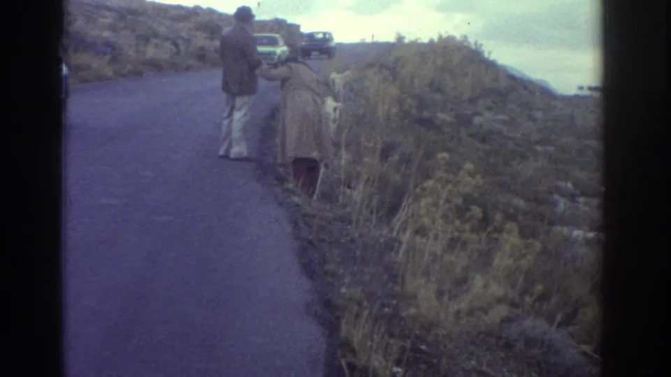 1976 Greece 8mm frame of two travelers standing on a roadside overlook, with parked cars ahead and scrubby hillside dropping toward the coast.