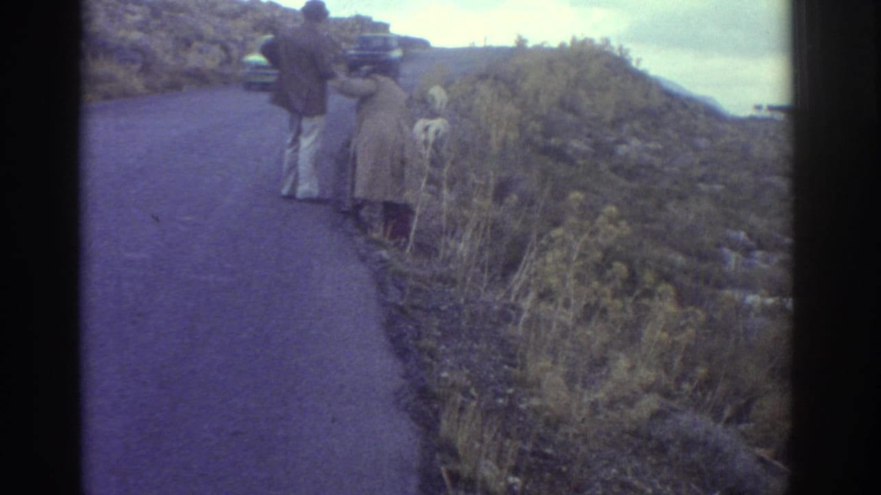 1976 Greece home-movie frame showing two adults on a winding mountain road near dry brush and a small white goat.