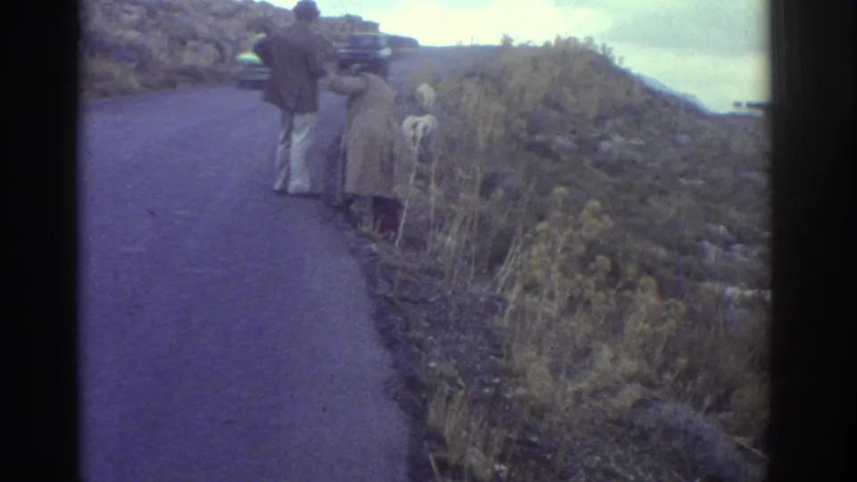 1976 Greece home-movie frame showing two adults on a winding mountain road near dry brush and a small white goat.