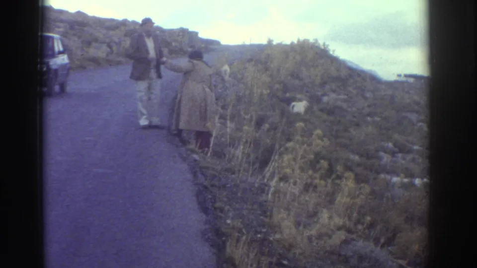 Home-movie frame from Greece in 1976 showing adults beside a mountain road, a goat, and a parked vintage car.