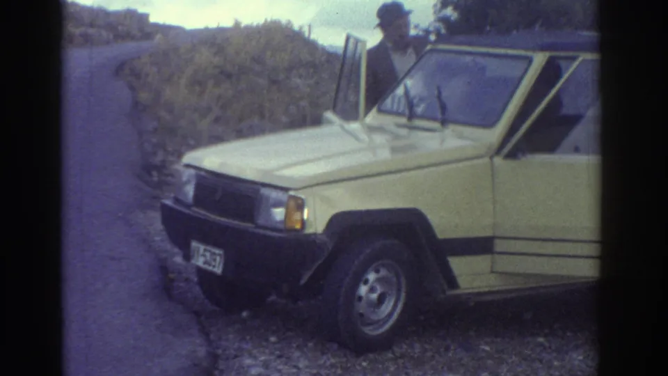 Close 1976 Greece home-movie view of a light yellow Renault-style compact car on a roadside shoulder, with a person nearby.