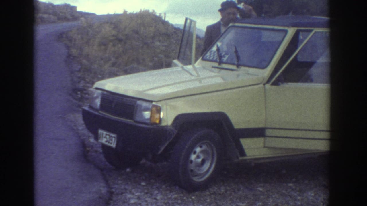 1976 Greece home-movie frame showing a man beside a yellow Renault 4L-style soft-top car parked on gravel by a coastal road.
