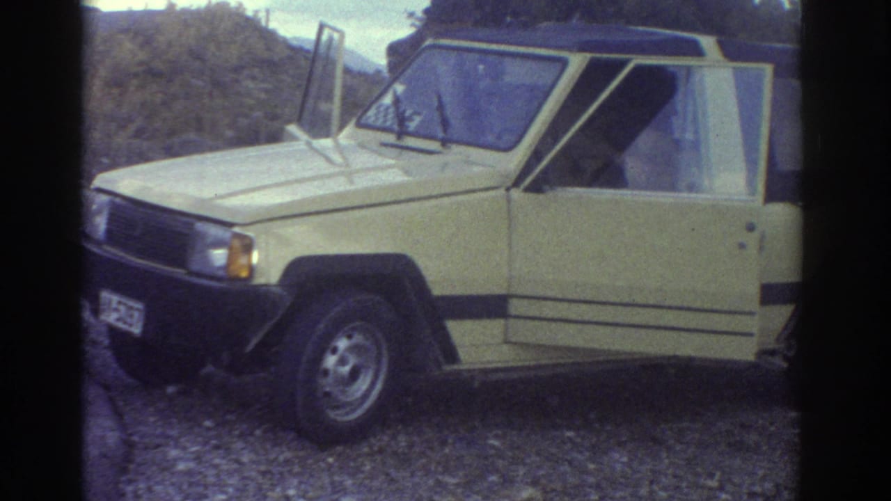 Yellow soft-top car with doors open sits on gravel beside a road and low coastal vegetation.