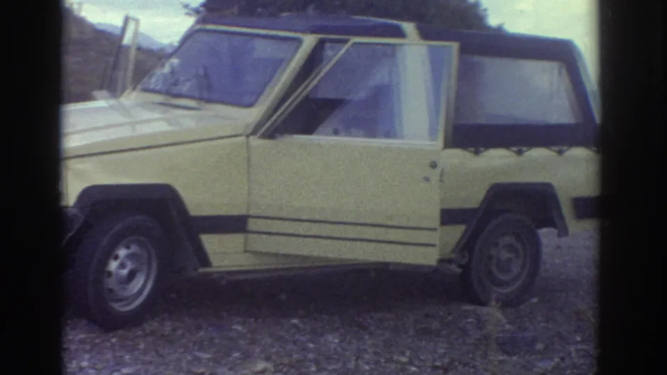 Side view of the yellow soft-top car in 1976 Greece, showing its open door, black roof, and roadside gravel setting.