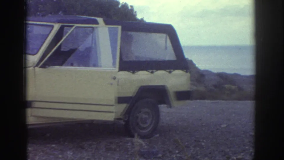 The car is framed from the rear side near the Greek shoreline, with the ocean visible beyond the gravel pull-off.