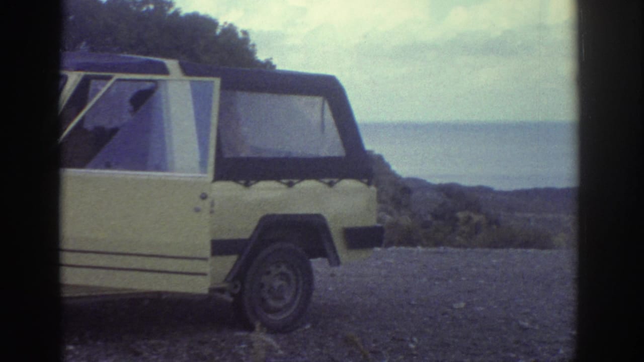 1976 Greece 8mm frame of a yellow open car at a gravel coastal overlook, with the sea and distant rocky shore in view.