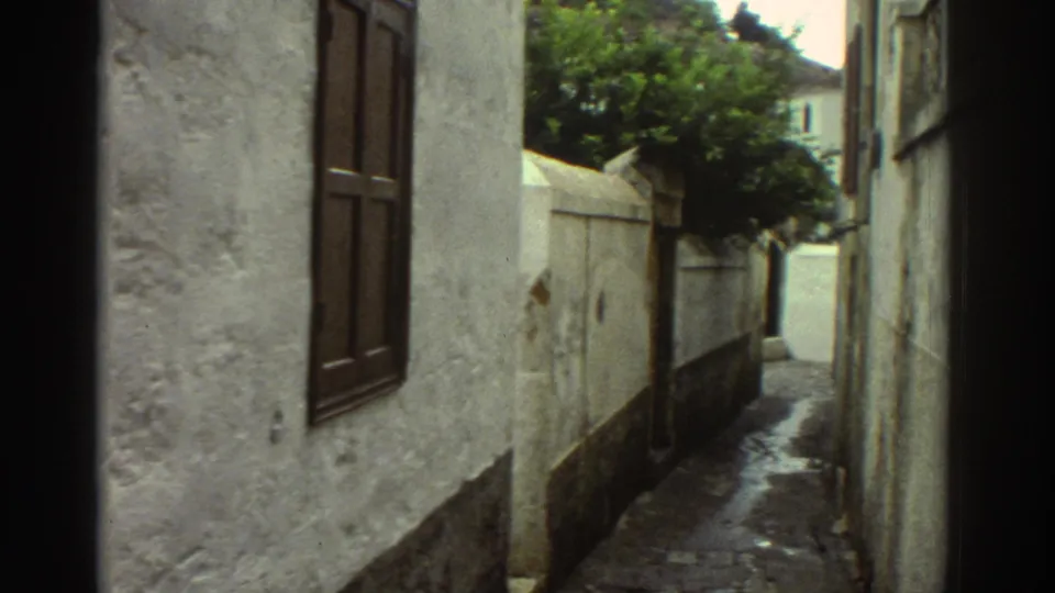 Handheld 8mm view of a Greek village passage in 1976, showing white walls, wooden shutters, cobblestones and a tree over the lane.