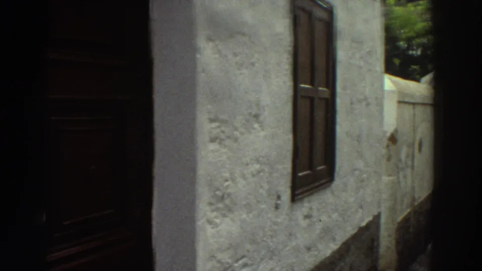 Close 1976 Greece 8mm frame of a white stucco building wall with a dark wooden shutter along a narrow village street.