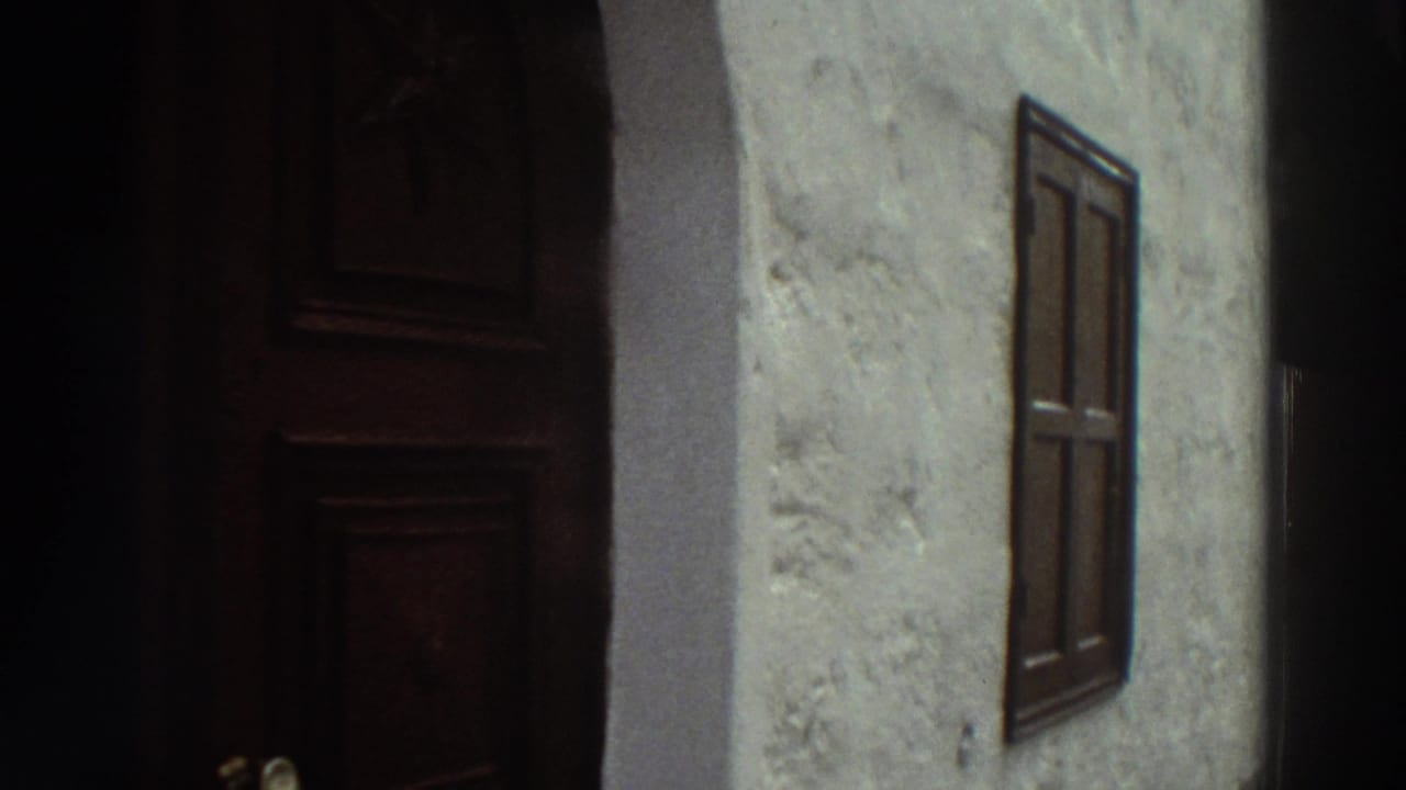 1976 Greece 8mm frame showing a whitewashed exterior wall, dark wooden door, small shuttered window, and shaded alley architecture.