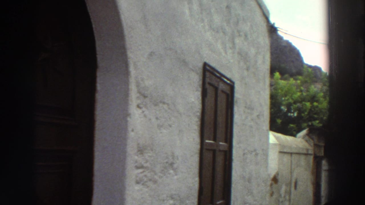 1976 Greece 8mm frame with a white stucco building, dark arched doorway, wooden shutters, green foliage, and rocky hillside beyond.