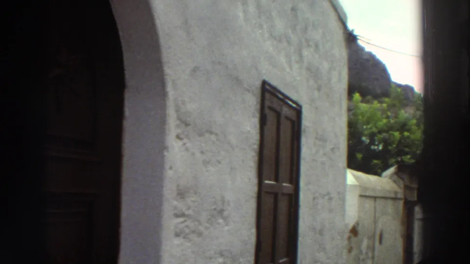 1976 Greece 8mm frame with a white stucco building, dark arched doorway, wooden shutters, green foliage, and rocky hillside beyond.