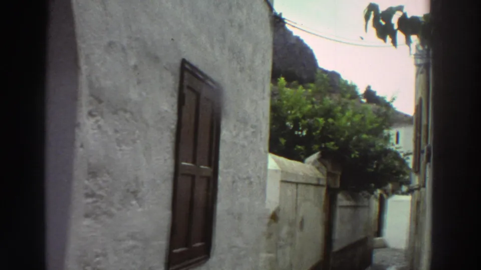 1976 Greece 8mm frame looking down a narrow alley past a whitewashed wall, shuttered window, trees, and a rocky hillside.