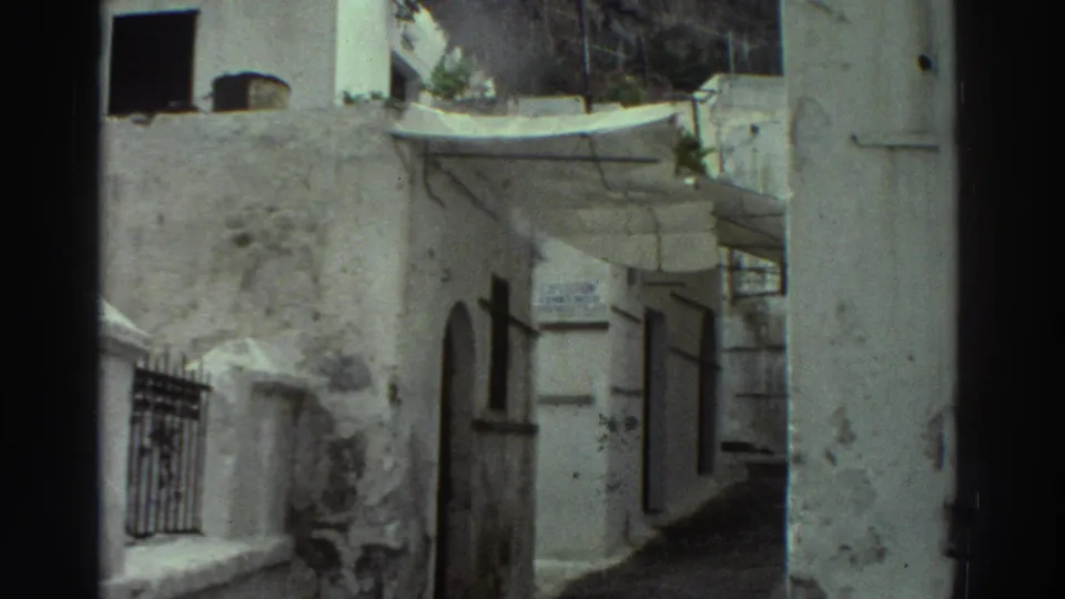 1976 Greece 8mm frame showing a whitewashed alley corner with arched openings, barred window, fabric awning, and doorway in shadow.
