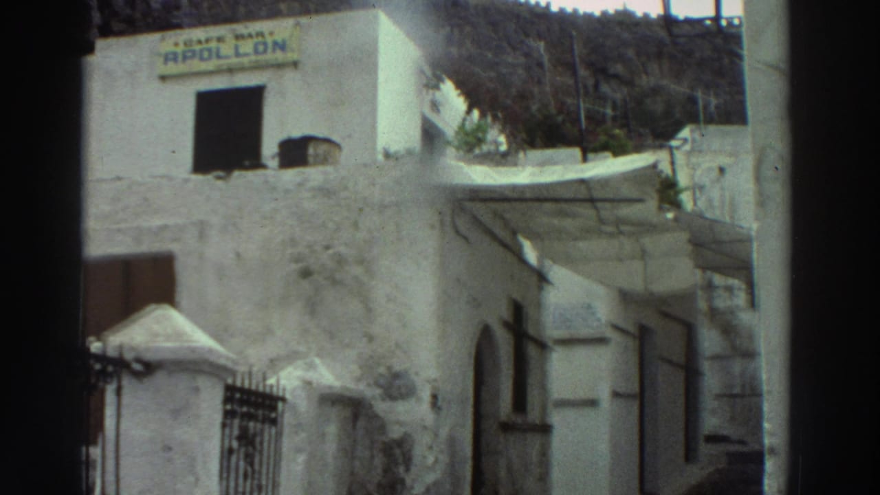 1976 Greece home-movie frame showing a whitewashed cafe building with a visible Cafe Bar Apollon sign and hillside behind it.