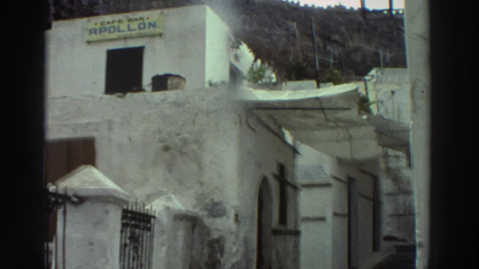 1976 Greece home-movie frame showing a whitewashed cafe building with a visible Cafe Bar Apollon sign and hillside behind it.