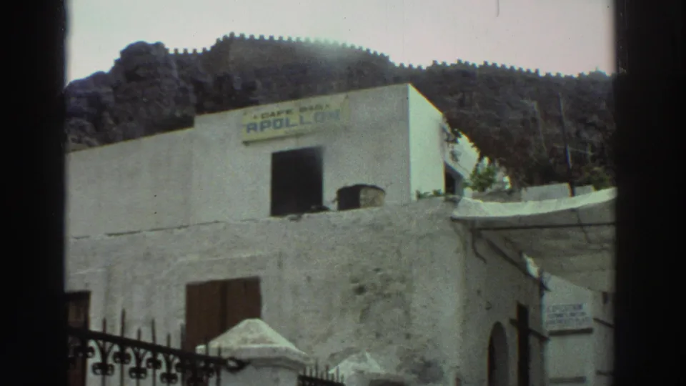 1976 Greece frame of a white-walled cafe facade with visible signage, dark window, gate detail, and rugged hillside in back.