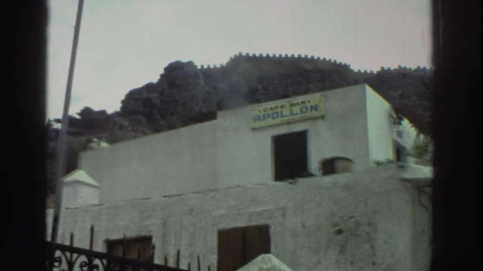 1976 Greece close framing of a whitewashed building marked Cafe Bar Apollon, with shutters, ironwork, and hillside beyond.