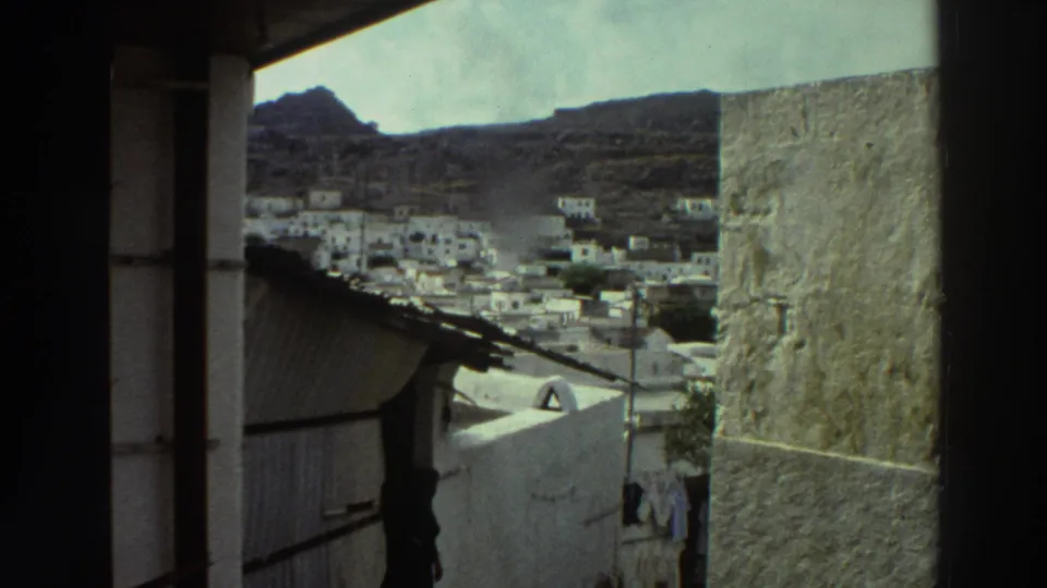 1976 Greece home-movie frame looking from a shaded side passage toward white village homes spread across a valley.
