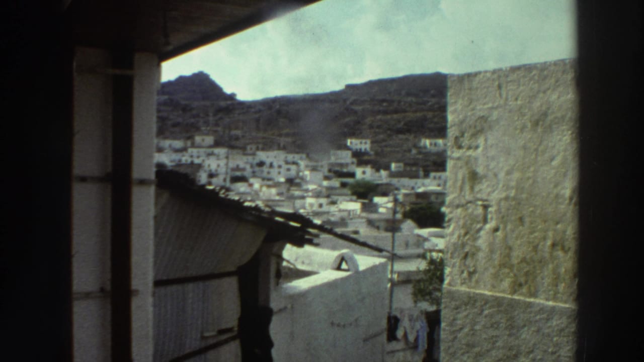 1976 Greece home-movie view of whitewashed hillside buildings framed by dark balcony posts and a shaded wall.