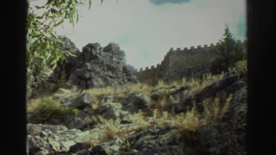 1976 Greece travel frame showing a rocky slope, dry vegetation, and battlement-like stone wall under bright sky.
