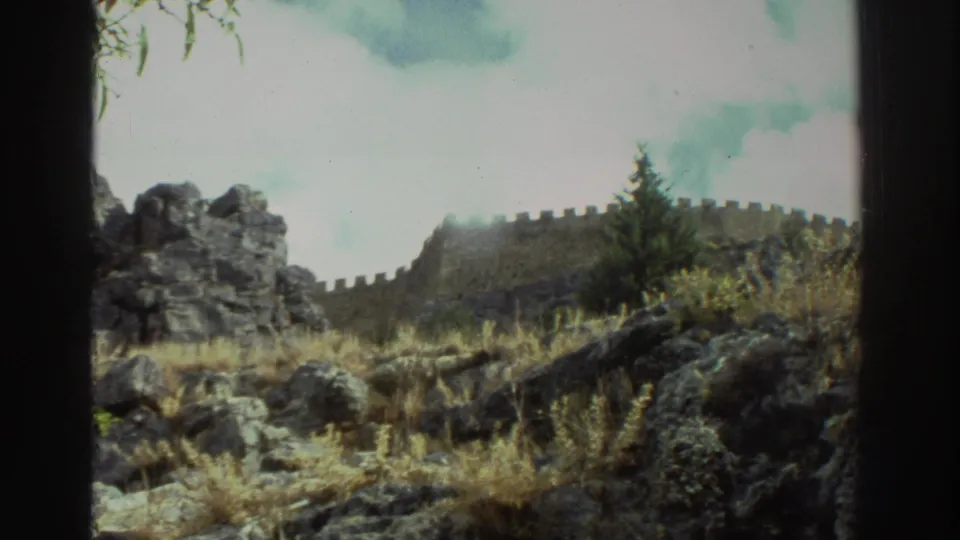 Rocky Mediterranean hillside in Greece with dry grass, a small evergreen, and a crenellated wall across the upper slope.