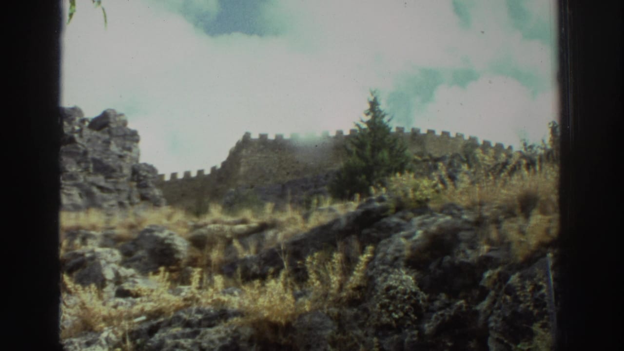1976 Greece 8mm frame showing dark rocks, dry vegetation, and an ancient-looking stone wall with battlements along a cliff edge.
