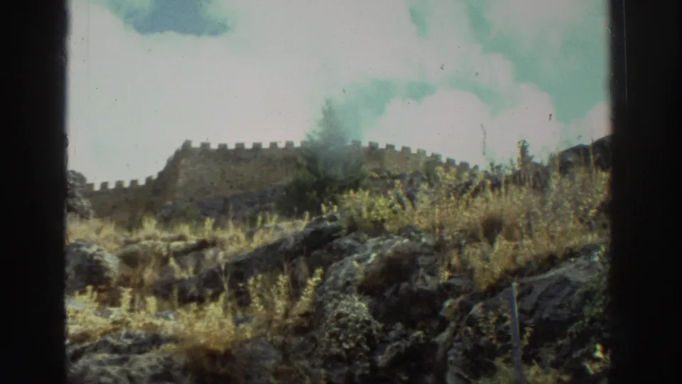 Vintage 8mm view in Greece of rocky ground, scrub, and a crenellated stone wall seen from a low visitor viewpoint.
