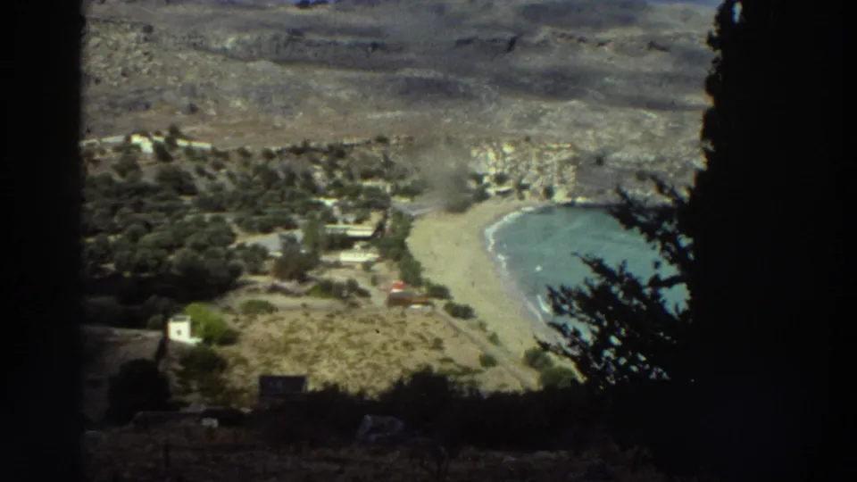 1976 Greece home-movie frame looking down from a shaded overlook toward a curved sandy beach, blue water, hillside, and small buildings.