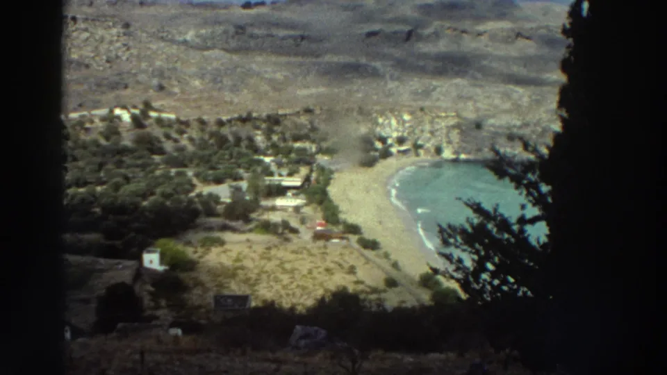 Vintage 8mm Greece frame showing a high view of a sheltered beach, blue-green water, hillside, and small coastal structures through dark foreground trees.