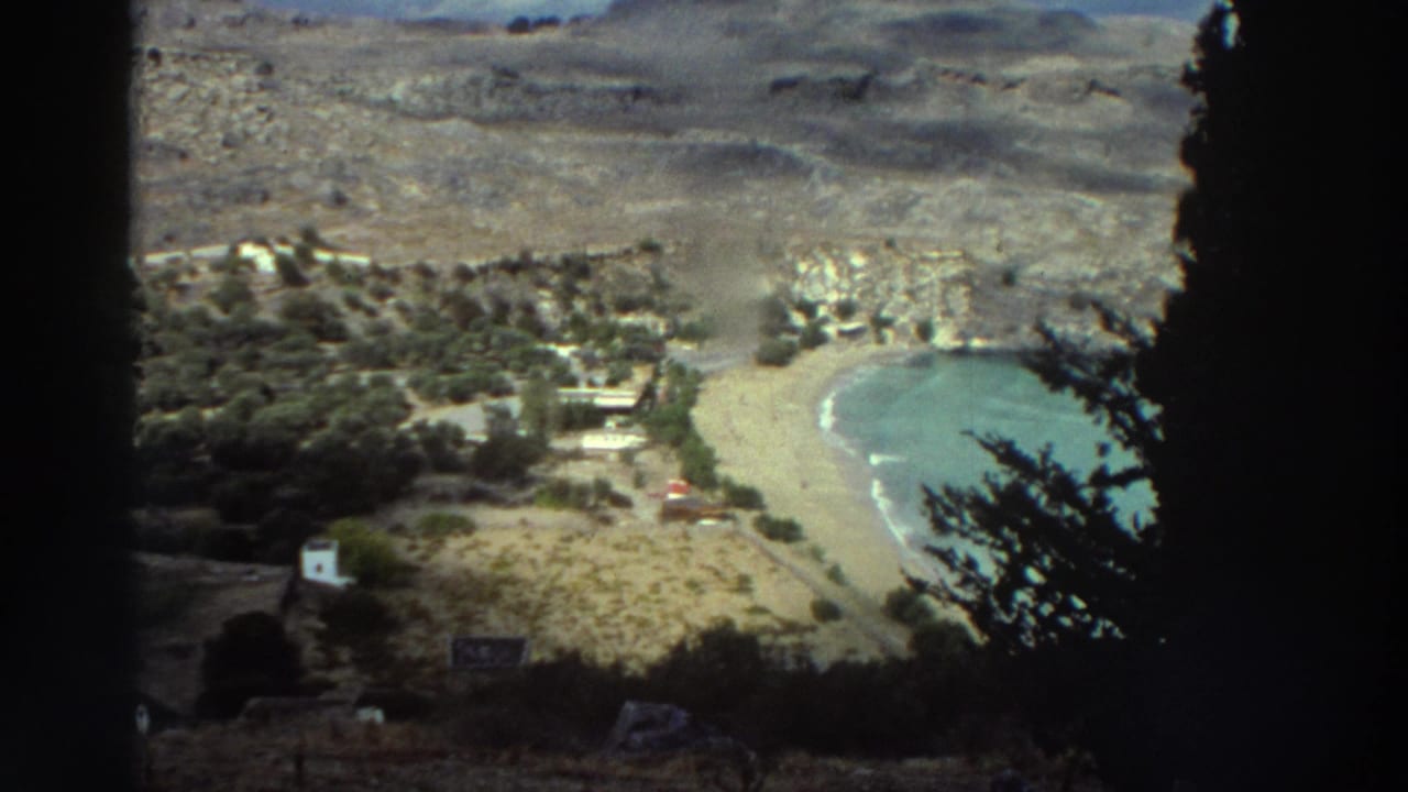 1976 Greece 8mm view looking down toward a curved sandy bay, rocky terrain, scattered buildings, and dark tree silhouettes.