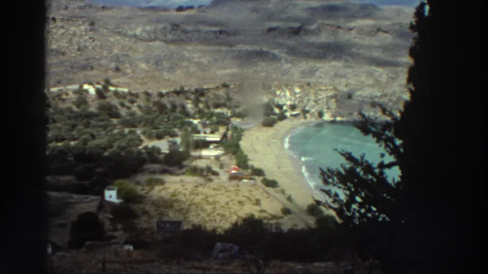 1976 Greece 8mm view looking down toward a curved sandy bay, rocky terrain, scattered buildings, and dark tree silhouettes.