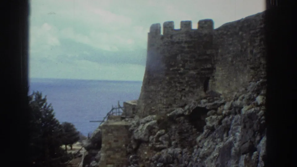 1976 Greece 8mm frame showing a crenellated stone castle wall above rocks, trees, and the blue sea beyond.