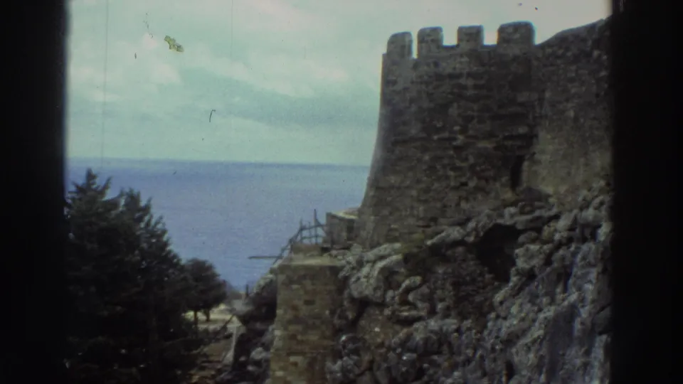 1976 Greece 8mm closer view of a weathered castle tower, stonework, rocky slope, cypress trees, and Aegean water beyond.