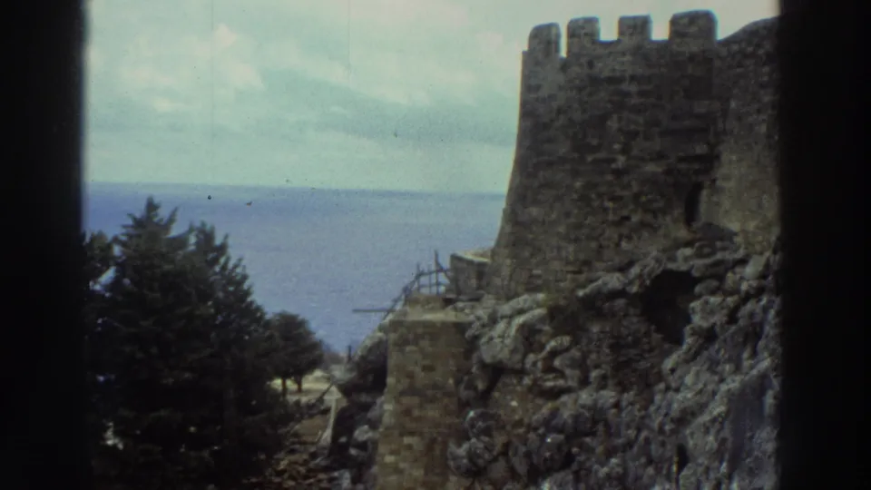 1976 Greece 8mm view of a coastal stone castle tower, rough rocks, trees, and open sea with dark vignetted frame edges.