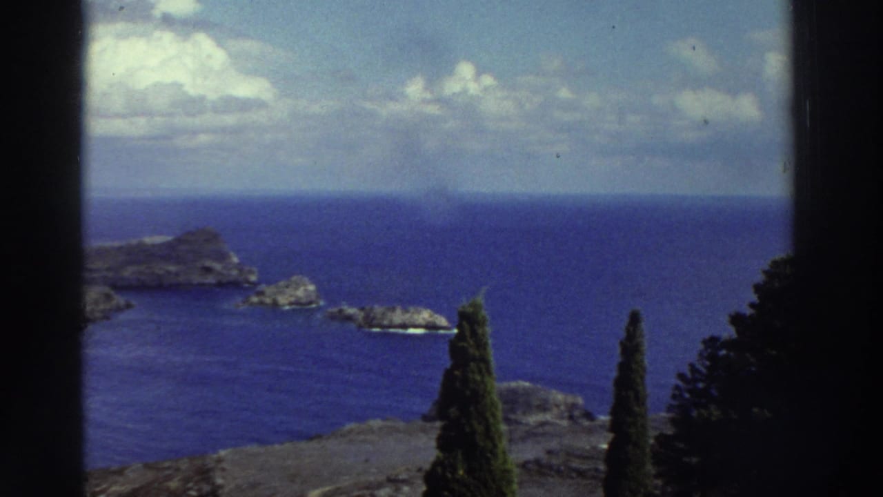 1976 Greece home-movie view of a rocky Mediterranean island, blue water, clouds, and dark cypress trees from an elevated overlook.