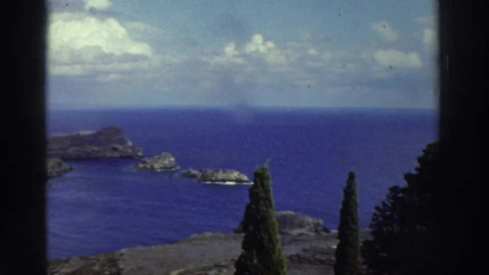 1976 Greece home-movie view of a rocky Mediterranean island, blue water, clouds, and dark cypress trees from an elevated overlook.