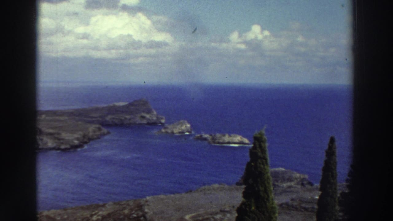 Rocky headland, offshore rocks, blue sea, clouds, and cypress trees within dark film borders.
