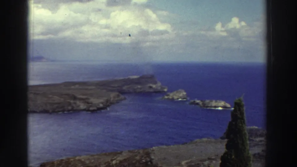 1976 Greece home-movie frame with a closer coastal view of a rocky peninsula, offshore rocks, blue sea, and a foreground cypress.