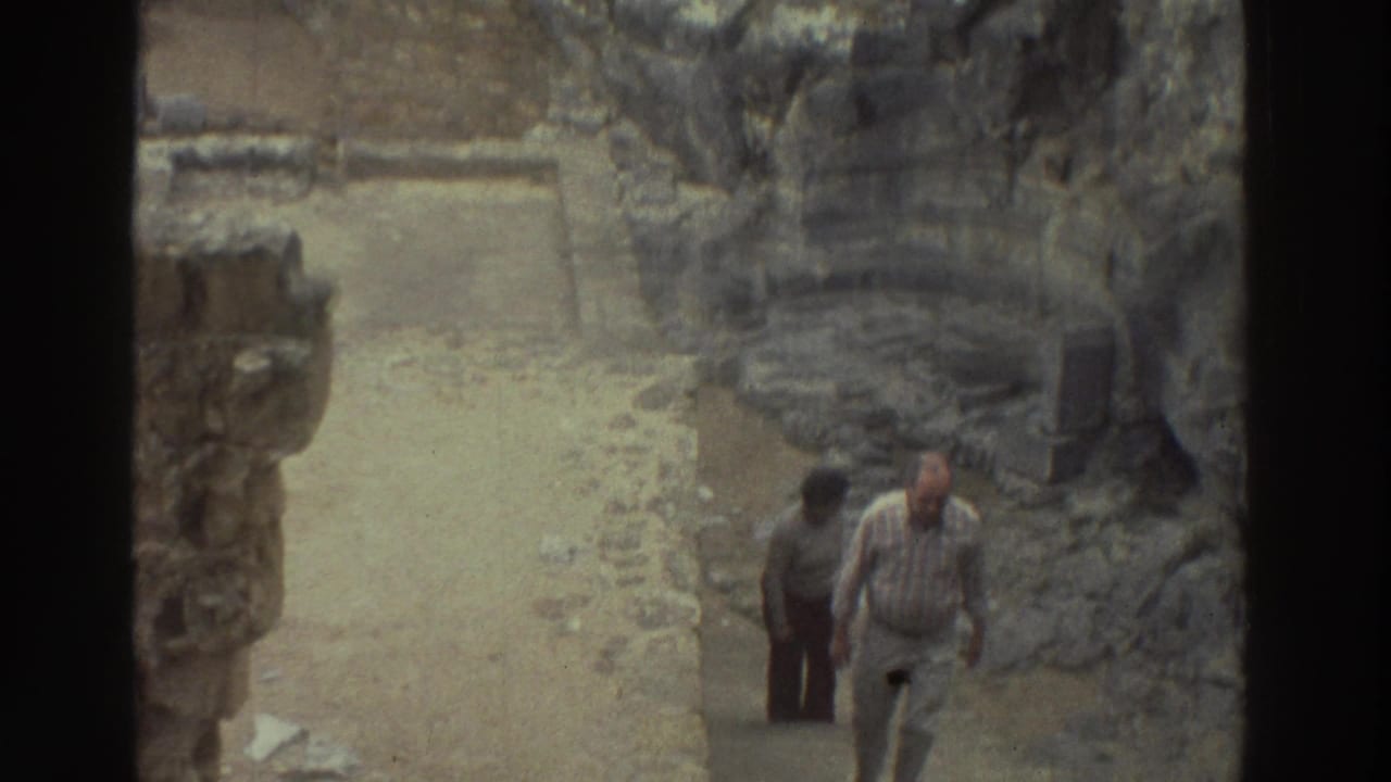 1976 Greece 8mm frame with two adult visitors walking through ancient stone ruins along a narrow path.