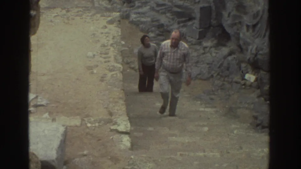 1976 Greece home-movie frame of two adult visitors climbing stone steps between weathered ruin walls.