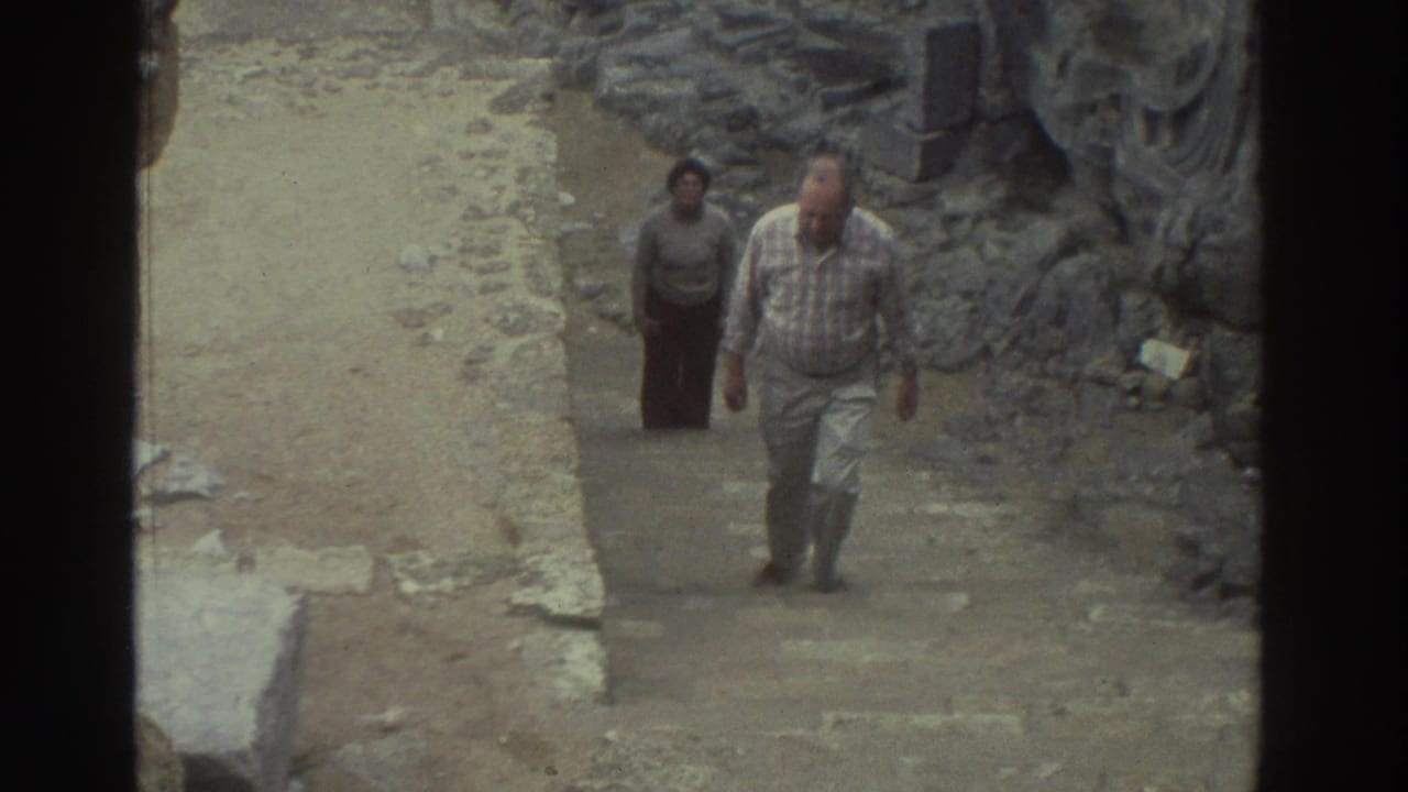 In 1976 Greece, a man and woman walk uphill through weathered stone ruins, seen through a dark-framed home-movie view.