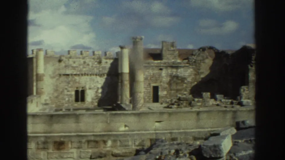The 8mm frame looks across weathered Greek ruin walls with columns, broken stonework, and foreground rubble.