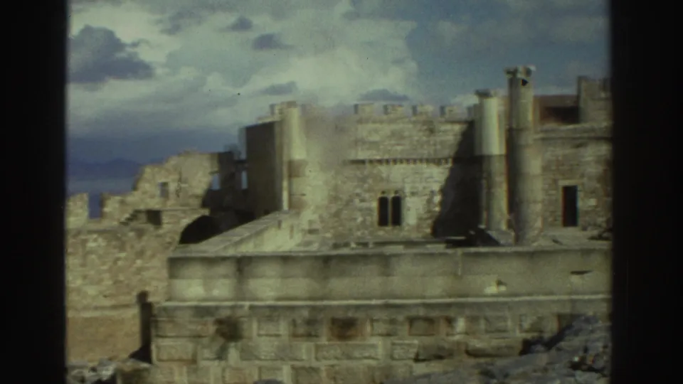 A 1976 Greece home-movie frame shows a stone ruin with upright columns, wall openings, and scattered rubble under a cloudy sky.