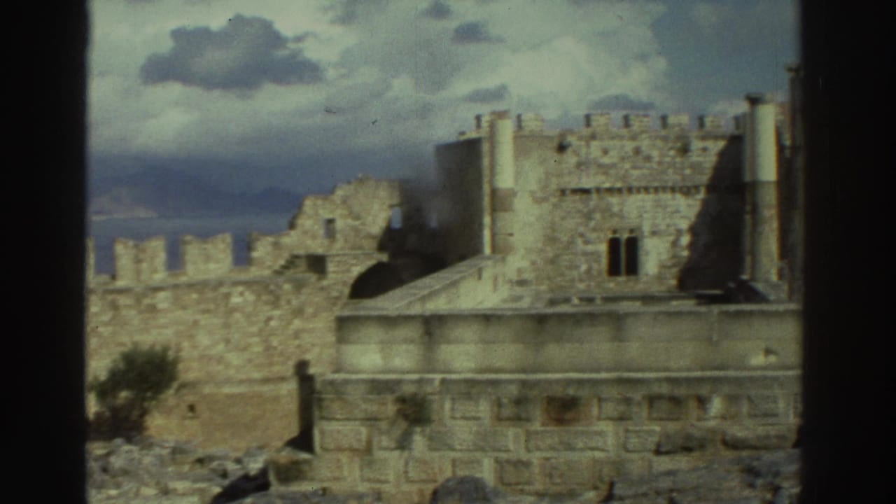 1976 Greece 8mm frame showing crenellated stone fort walls, ruined towers, rocky ground, and cloudy coastal sky.